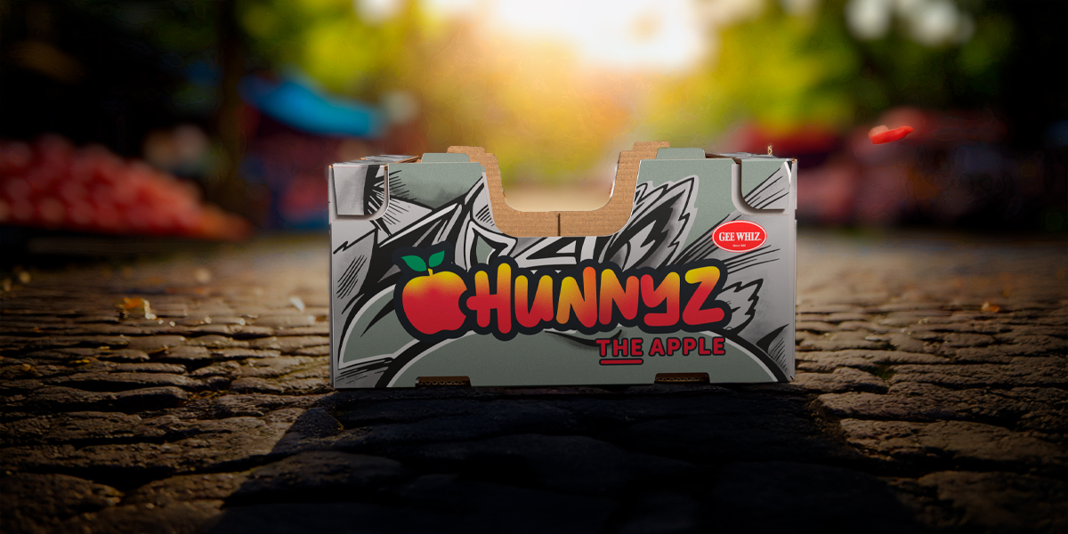 Hunnyz apple box by Gee Whiz with bold, colorful branding and an illustrated apple logo, placed on a cobblestone street with a bright, blurred outdoor market scene in the background.